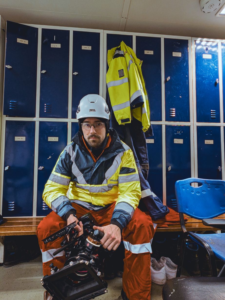Kameramann mit Filmkamera, Helm und Schutzkleidung an Bord eines Offshore-Schiffs